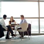 Two sales professionals and a client are engaged in a deep discussion around a table in a bright office space, illustrating the technique of creating customer needs through questioning.