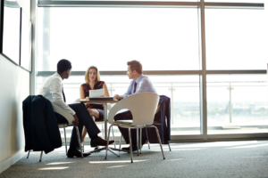 Two sales professionals and a client are engaged in a deep discussion around a table in a bright office space, illustrating the technique of creating customer needs through questioning.