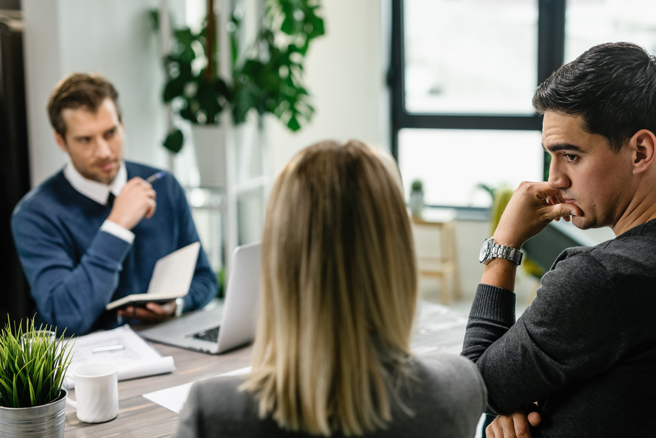 A salesman in a modern office setting attentively listens to two clients who sit across from him. This scene illustrates techniques to overcome sales stalls, with the salesman appearing to thoughtfully consider the clients' conversation.