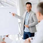 A smiling salesman points to a flip chart, set to deliver a them-centric demo as a winning sales strategy to an engaged audience.