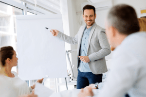 A smiling salesman points to a flip chart, set to deliver a them-centric demo as a winning sales strategy to an engaged audience.