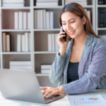 A saleswoman is smiling while talking on the phone and working on her laptop, exemplifying the strategy of choosing phone meetings over phone tag for sales success.