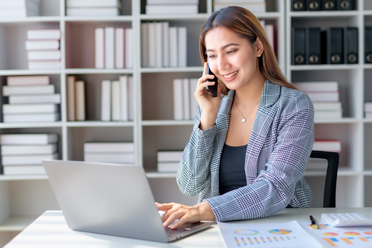 A saleswoman is smiling while talking on the phone and working on her laptop, exemplifying the strategy of choosing phone meetings over phone tag for sales success.